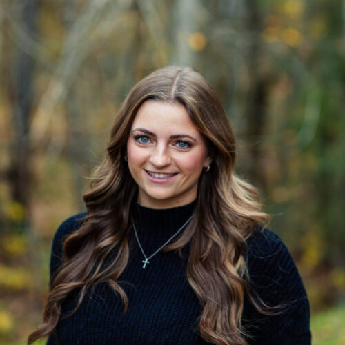 A woman with long, wavy brown hair wearing a black sweater and a necklace with a cross pendant smiles outdoors in a blurred, wooded background.