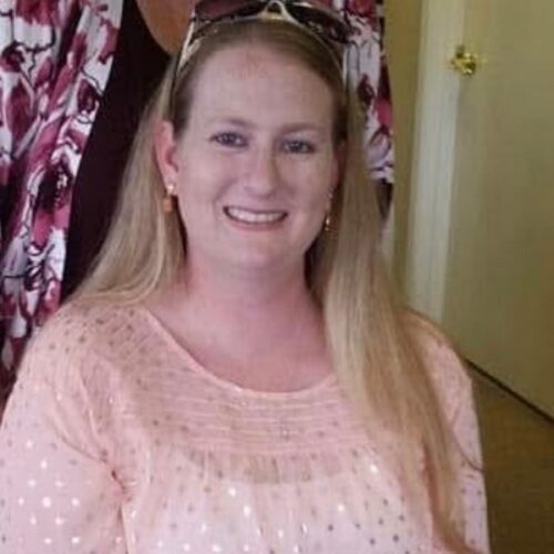 A woman with long blonde hair, wearing a light pink blouse with a dotted pattern and sunglasses on her head, smiles while sitting indoors. A floral-patterned garment is visible behind her.