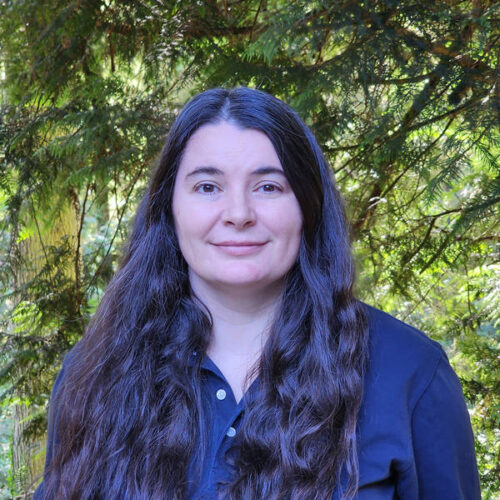 A woman with long, dark wavy hair and a navy blue shirt smiles softly, standing outdoors in front of green, leafy trees. Sunlight filters through the branches in the background.