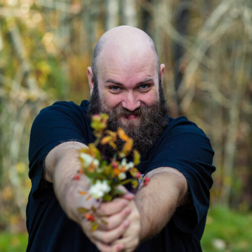 A bearded man with a shaved head smiles mischievously while holding out a small bouquet of wildflowers toward the camera, standing outdoors with blurred trees in the background.