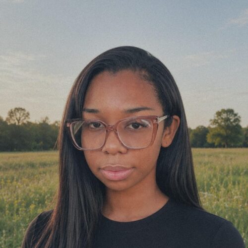 A young woman with long straight hair and large clear glasses stands in a grassy field with trees in the background, under a blue sky with light clouds. She is wearing a black shirt and looking at the camera with a neutral expression.