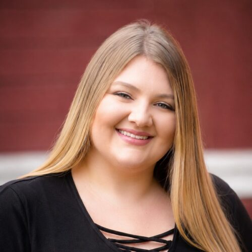 A young woman with long, straight blonde hair smiles at the camera. She is wearing a black top with crisscross detail at the neckline. The background is blurred and red in color.