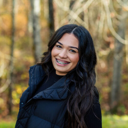 A young woman with long dark hair and braces smiles outdoors, wearing a black puffer vest and black top. The background is blurred with trees and autumn colors.